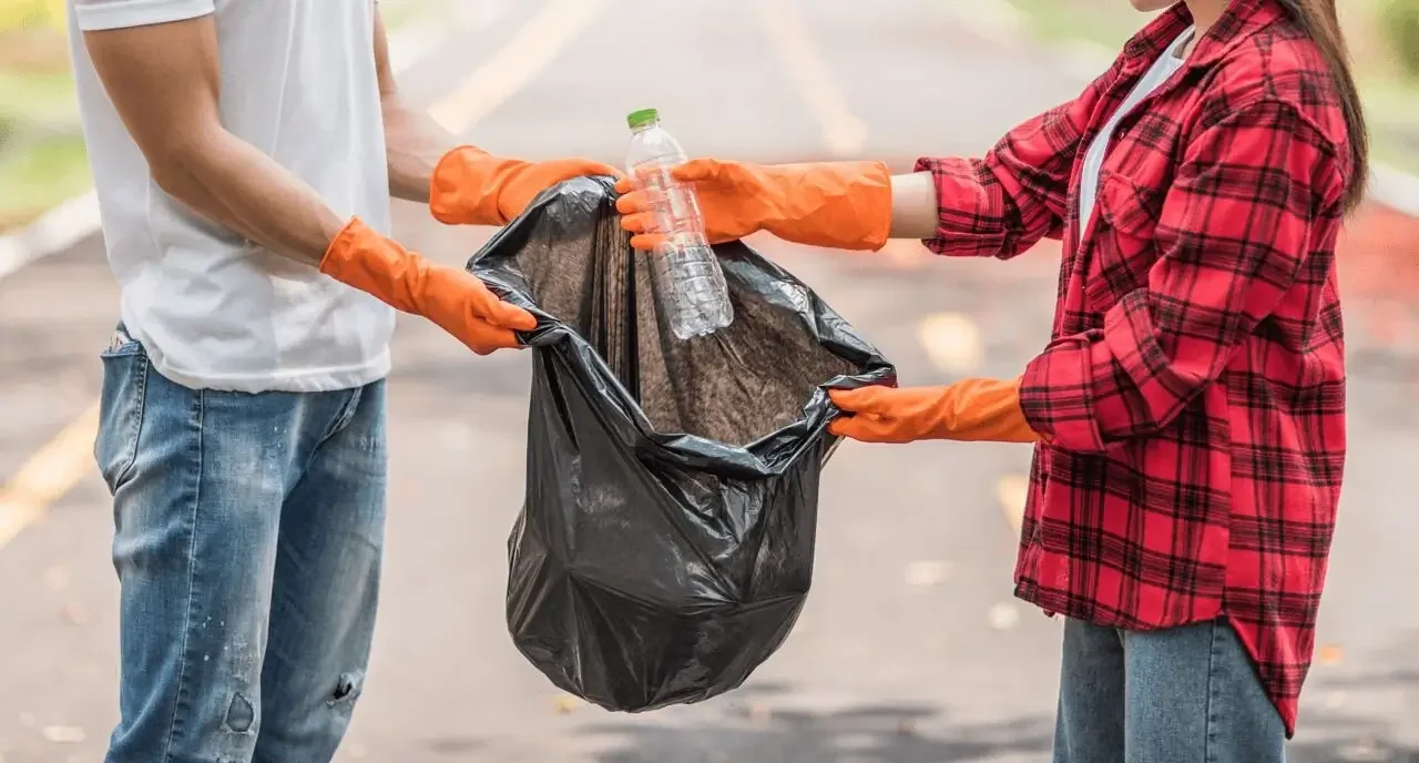 Zwei Personen mit orangen Handschuhen sammeln Müll, eine hält eine schwarze Tüte, die andere reicht eine Plastikflasche hinein.
