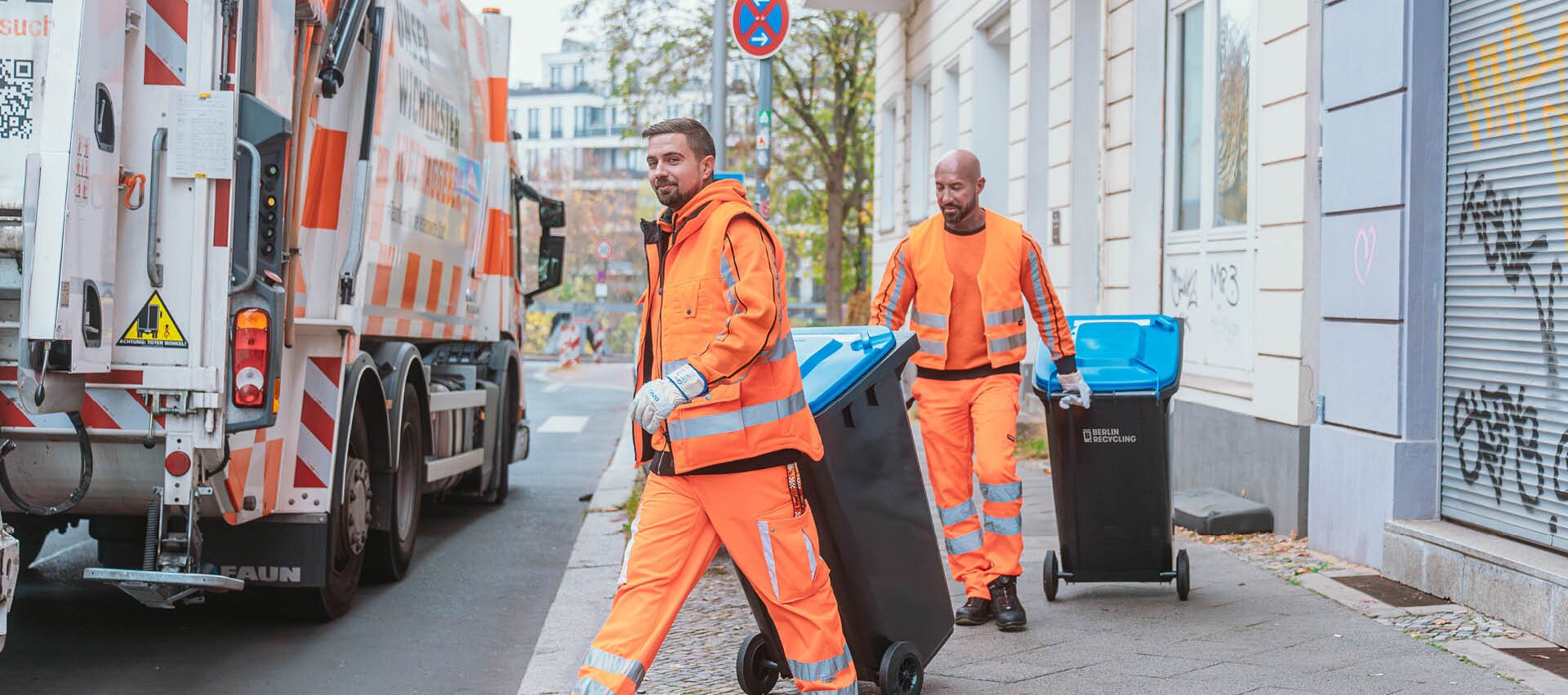 Zwei Müllwerker in orangefarbener Schutzkleidung schieben Mülltonnen auf einem Bürgersteig neben einem Müllfahrzeug in einer städtischen Straße.