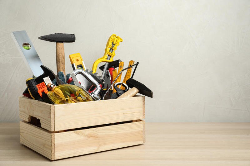 Holzkiste mit verschiedenen Handwerkzeugen wie Hammer, Wasserwaage, Schraubenzieher und Schutzbrille auf einem Tisch