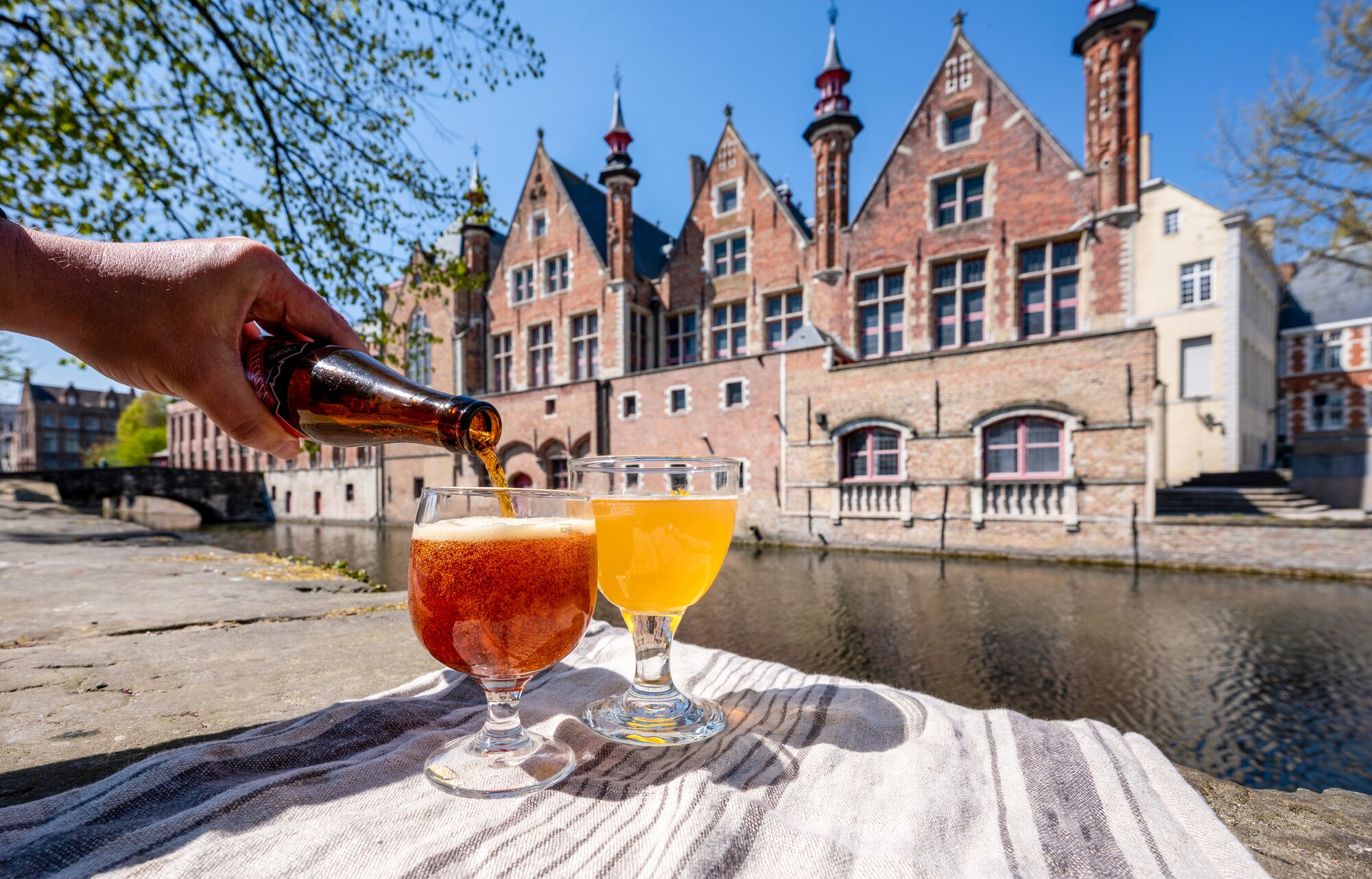 An arm pouring a beer into a glass next to another glass of beer in front of a river and buildings in Bruges, Germany