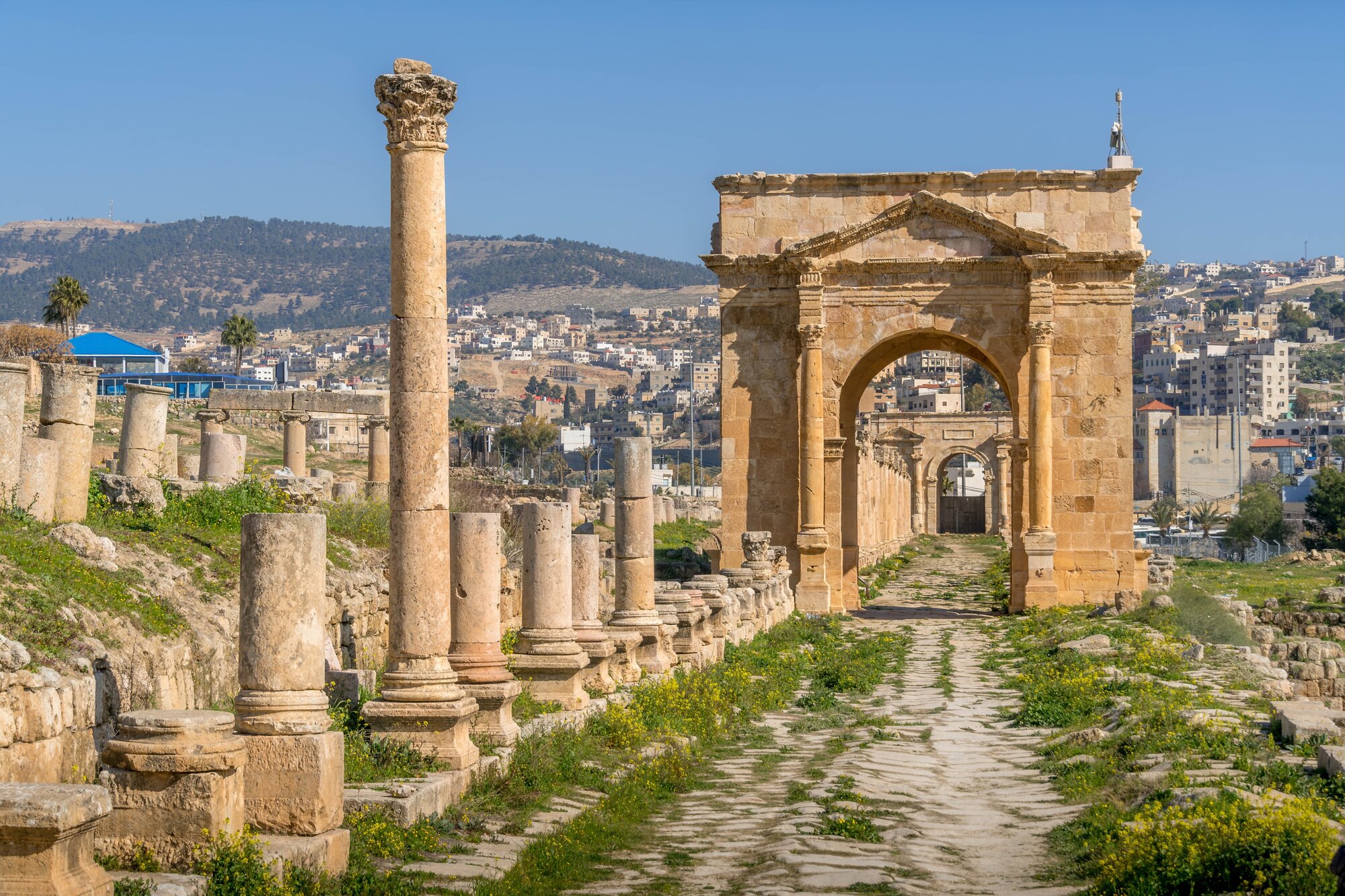 Historic Roman ruins in Jerash, Jordan, showcasing a colonnaded street and grand arch, set against rolling hills and a modern cityscape.