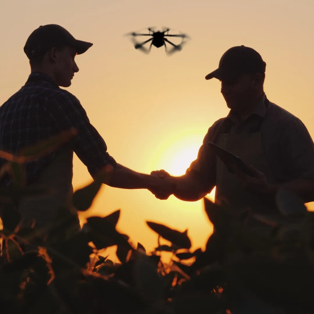 Two people shaking hands in a field at sunset with a drone above them