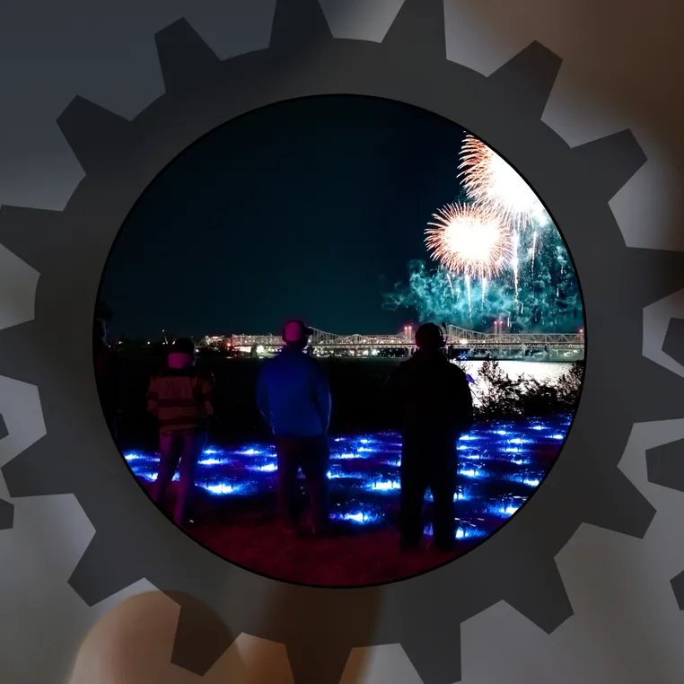 Silhouetted people watching a fireworks display over a bridge at night, with blue LED ground lights illuminating the area, framed within a gear-shaped overlay.