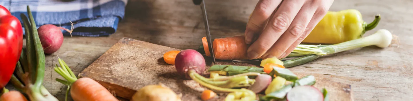  A close-up, panoramic shot of a person's hand using a knife to chop a carrot on a wooden cutting board. A variety of other vegetables, including radishes, green onions, and bell peppers, are visible on and around the board. A blue and white checkered towel is visible in the background. The lighting is natural and bright, creating a rustic and healthy feel.