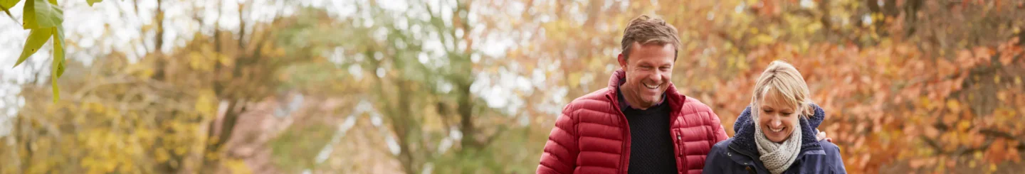  A panoramic shot of a smiling middle-aged man and woman walking together in a park during autumn. The man is wearing a red puffer jacket and the woman is wearing a dark blue jacket and a scarf. The background is filled with blurred trees and foliage in various shades of yellow, orange, and green. The image conveys a sense of happiness, health, and outdoor activity.