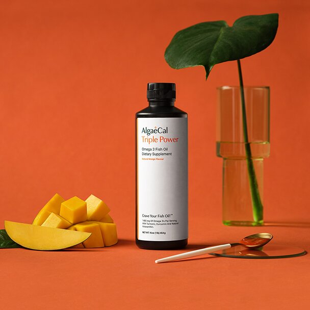 A studio product shot shows a bottle of AlgaeCal Triple Power Omega 3 Fish Oil. The scene also includes cut mango, a spoon with fish oil, and a glass vase with a leaf against an orange background.