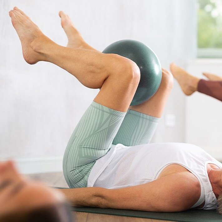 A person is doing a Pilates exercise with their legs raised, squeezing a small ball between their knees. 