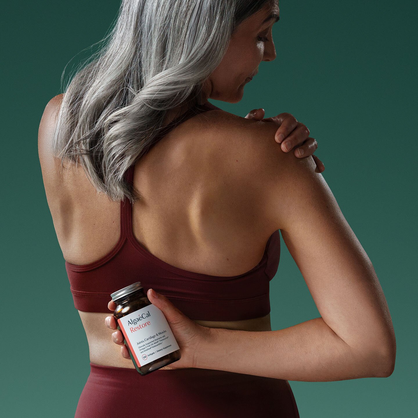 a woman wearing workout clothing, with medium length grey hair holds a bottle of AlgaeCal Restore behind her back