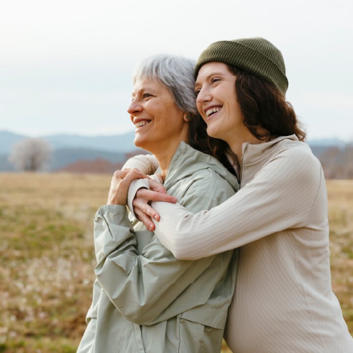A smiling daughter stands with her arms around her also smiling mother in an open field.