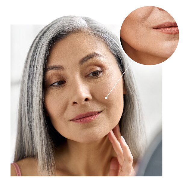  A middle-aged woman with long gray hair looks at her reflection in a mirror and touches her face. An inset circle shows a magnified, close-up view of her jawline and mouth, highlighting her smooth skin..