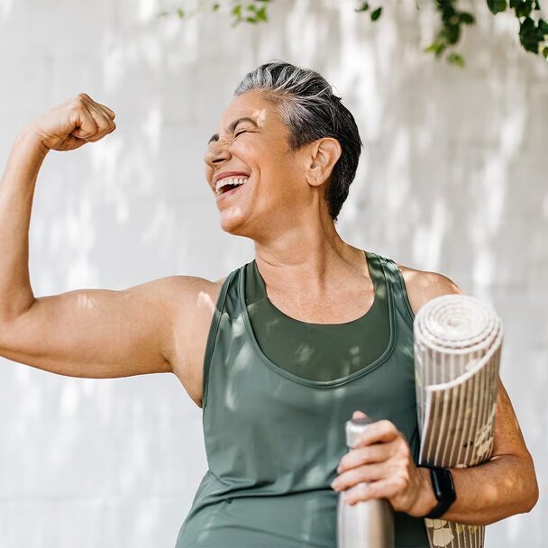  A joyful, gray-haired woman smiles with her eyes closed as she flexes her right arm in a bicep pose. She is holding a yoga mat under her left arm and a water bottle in her hand.