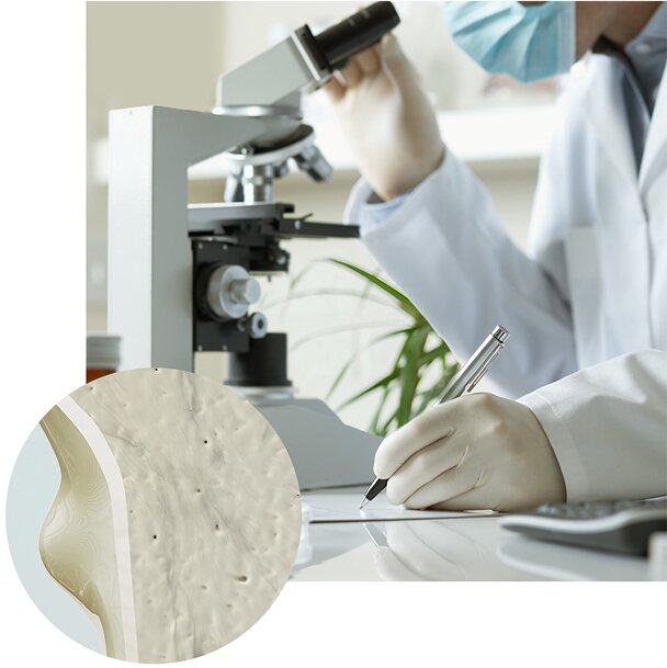  A lab scientist in a mask and white coat looks into a microscope and takes notes. An inset circle shows a close-up, digital rendering of porous bone tissue.