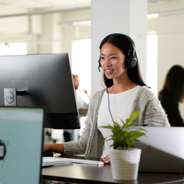 A young woman with a headset smiles as she works at a computer in a modern office.