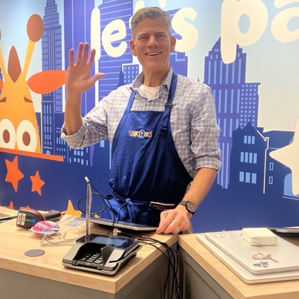 Friendly Go! Retail Group franchise operator waving and smiling in blue Toys R Us apron at store checkout counter with colorful branded wall behind