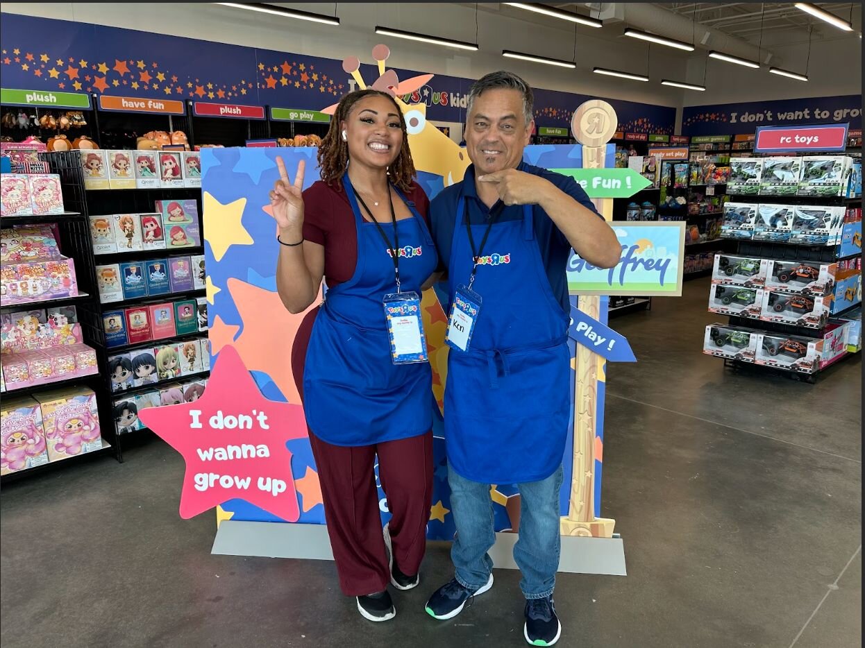 Two smiling Go! Retail Group store team members in blue aprons giving peace signs in a vibrant toy store filled with colorful products