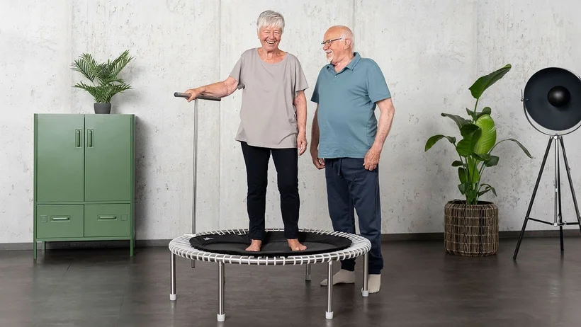 Une femme plus âgée s’entraîne sur le mini-trampoline tandis que son mari la soutient.