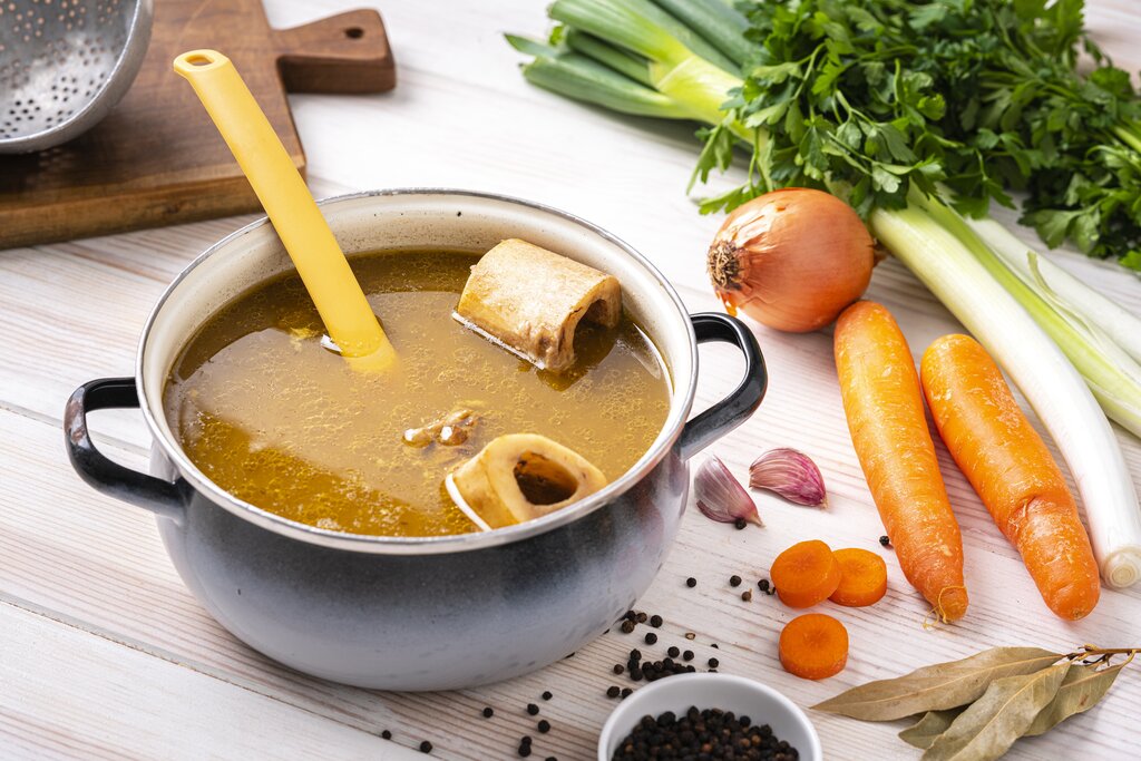 cooking pot with bone broth inside, surrounded by vegetables 