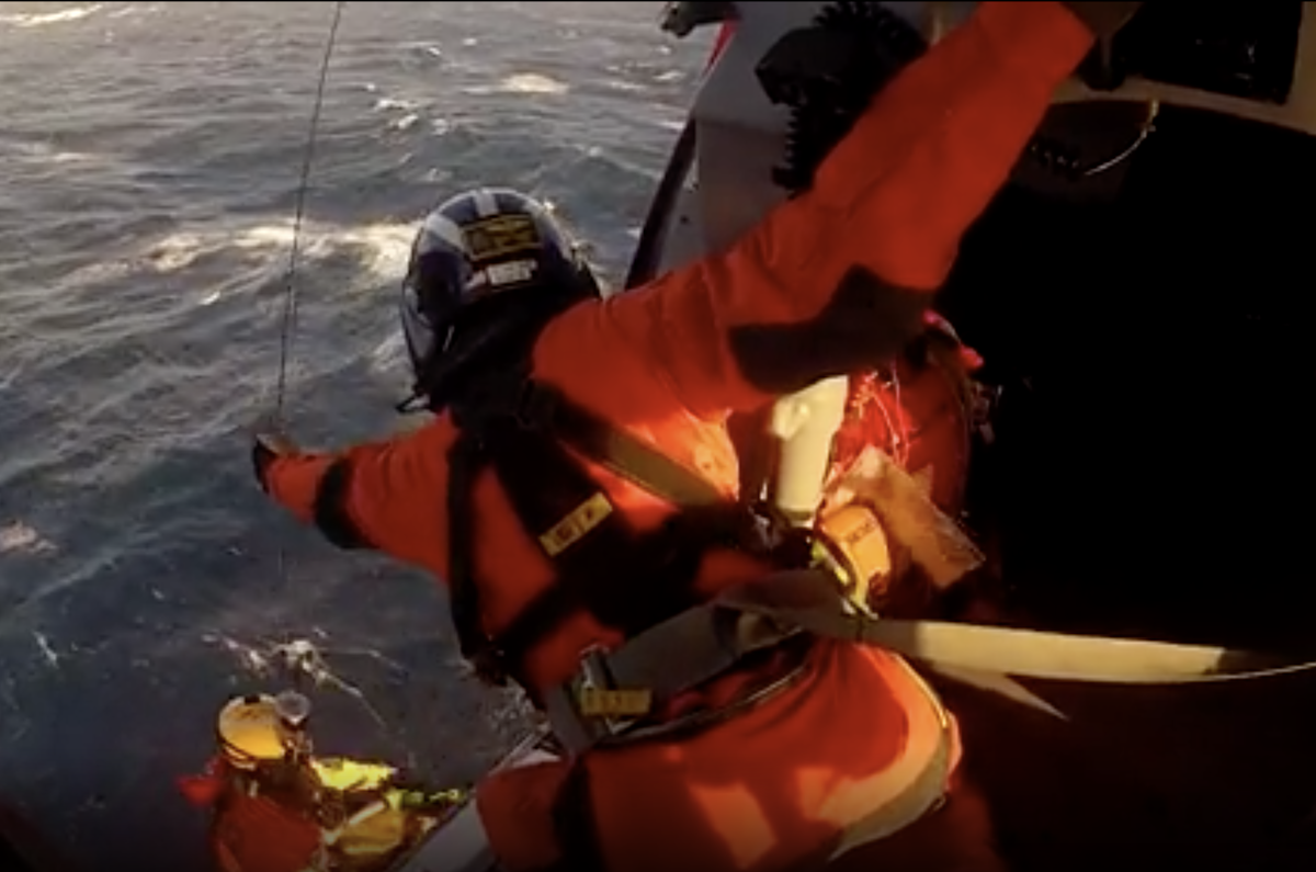 A Coast Guard flight engineer guides the hoist cable upwards as the a rescue swimmer nears the door of theri MH-65 helicopter during a rescue 80 miles off off shore of 6 people from a sinking sailboat. Coast Guard video.