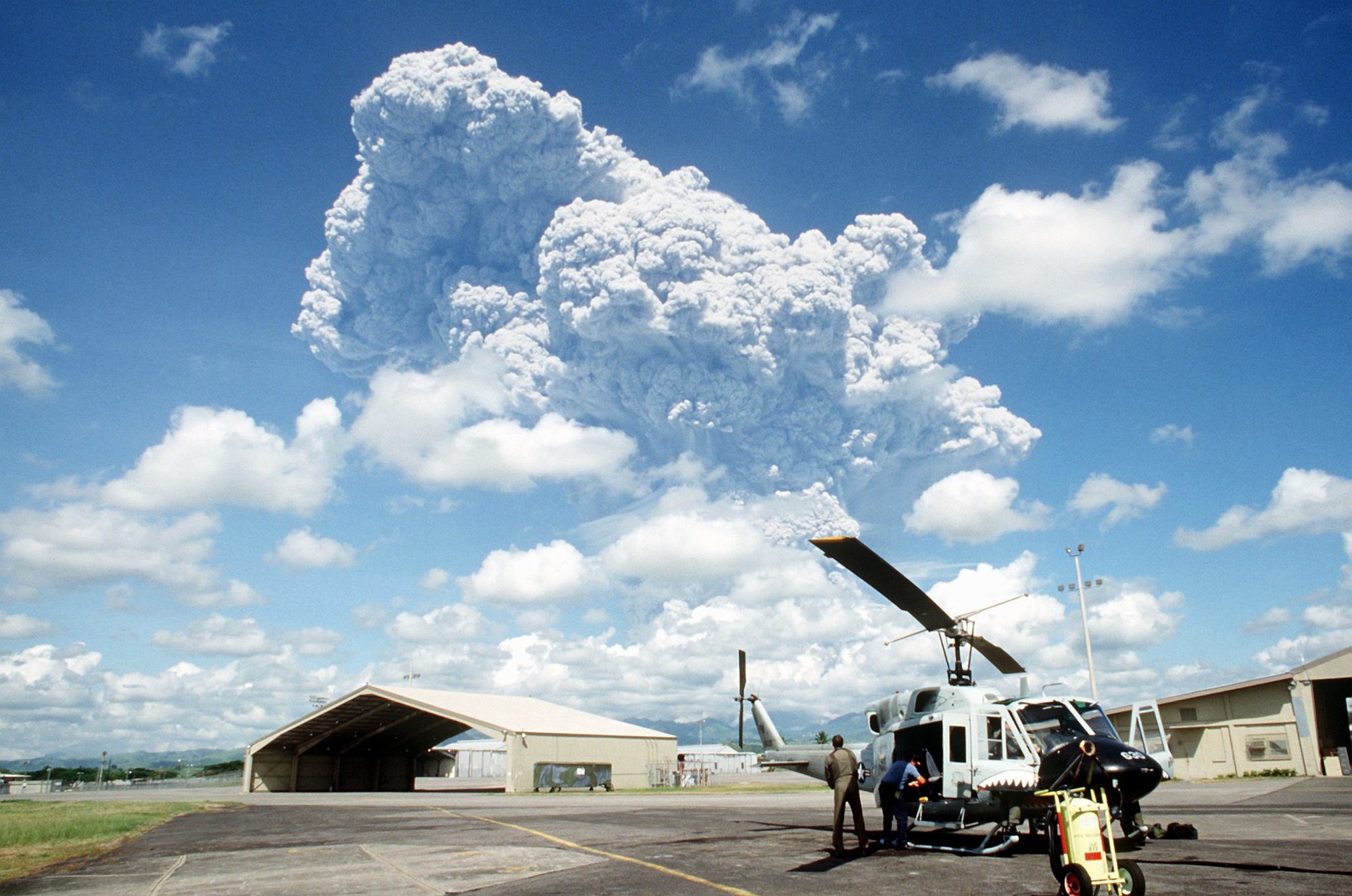 A helicopter crew member stands beside his UH-1N Iroquois helicopter as he watches a cloud of ash pour from Mount Pinatubo, a volcano that came alive for the first time in over 600 years.  Photo via U.S. National Archives.