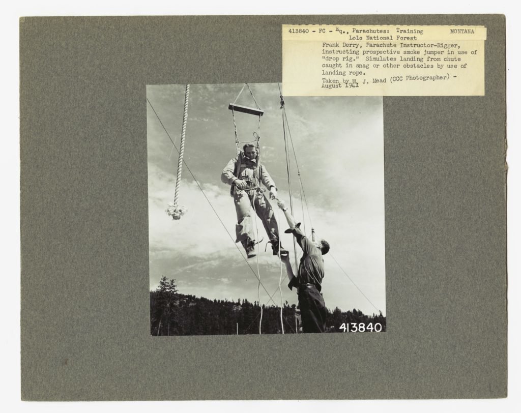 Frank Derry, Parachute Instructor-Rigger, instructing prospective smoke jumper in the use of the "drop rig." Simulates landing from chute caught in a snag or other obstacles by use of landing rope. Lolo National Forest, Montana. Photo by W.J. Mead, courtesy fo the National Archives and Records Administration.