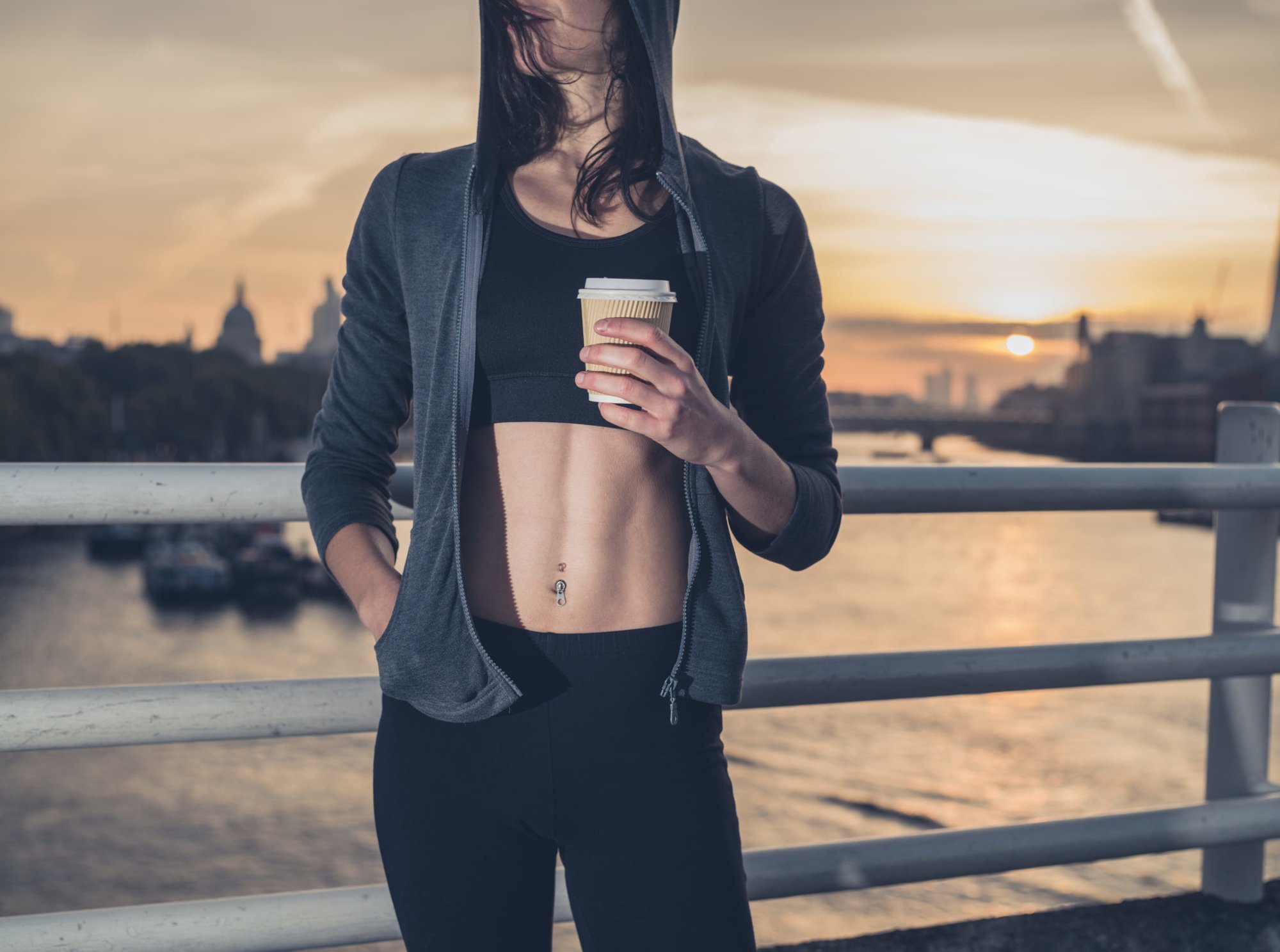 An athletic young woman with toned abs  is standing on a bridge in London at sunrise with a paper cup in her hand