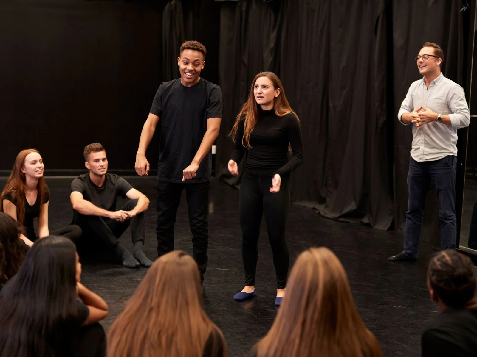 Group of people participating in an improvisation exercise, with two individuals performing in front of a seated audience, demonstrating teamwork and spontaneous collaboration.