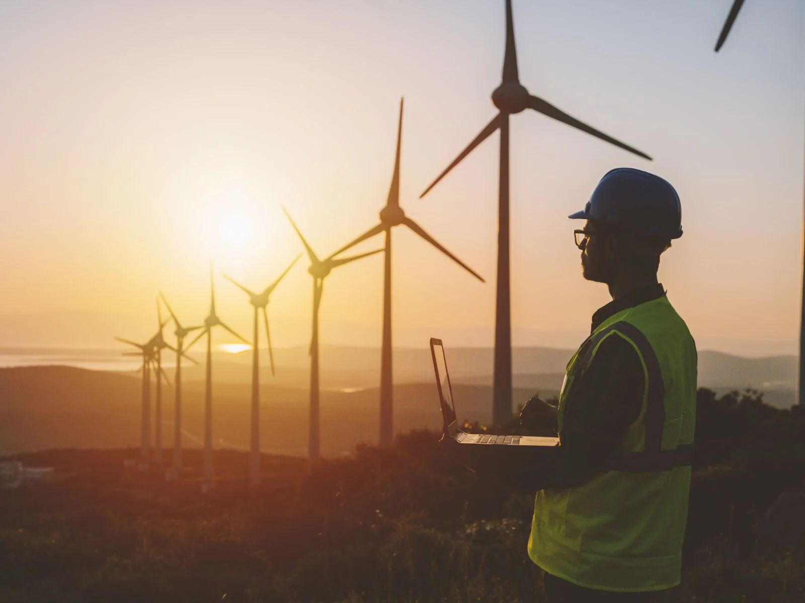 A renewable energy engineer in a hard hat and safety vest working on a laptop at a wind farm during sunset, symbolizing digital transformation in the energy sector