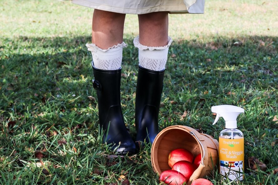 Girl's feet in black rain boots standing next to a basket of apples and Truly Free Home Fruit & Veggie Wash Spray.