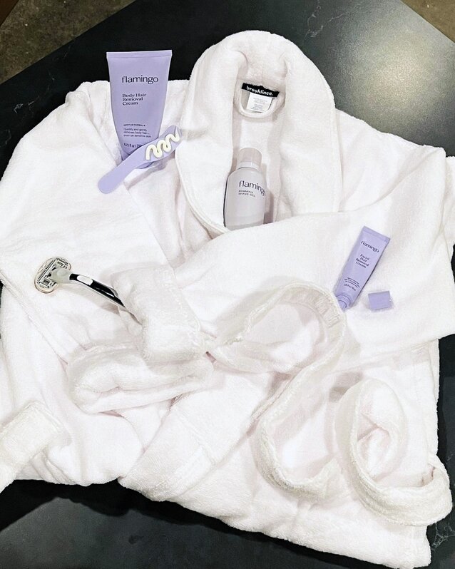 Soft white cotton robe with Flamingo hair removal products scattered on top of it including the Chrome razor, shave gel, and hair removal cream