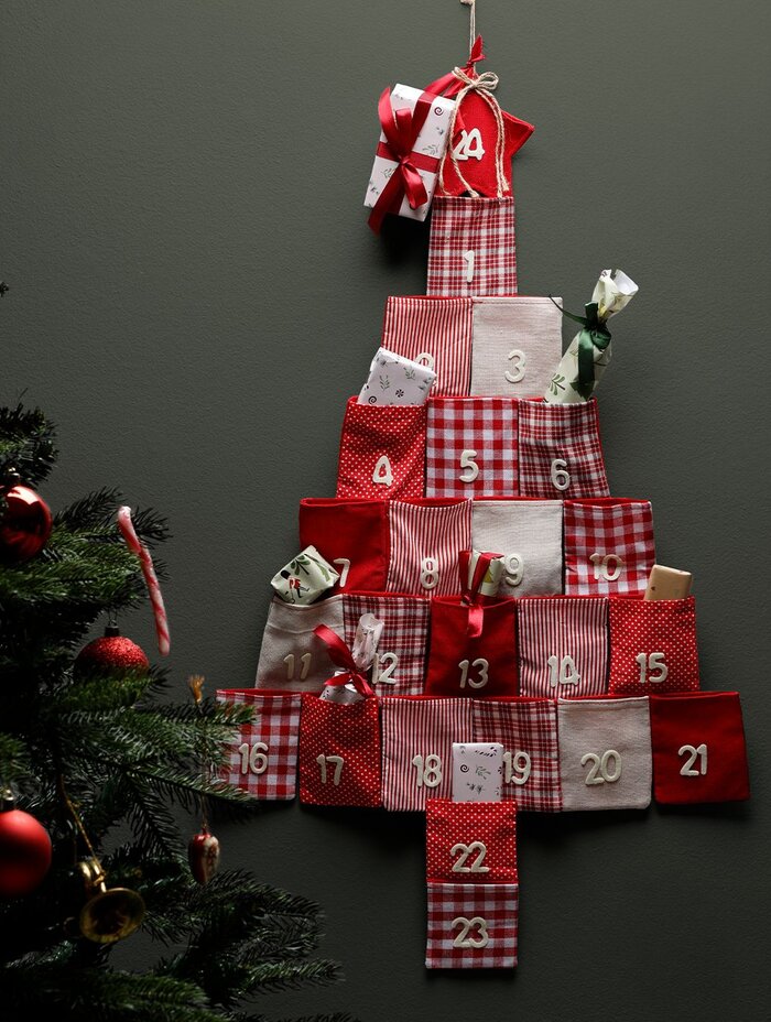 Julekalender formet som et juletræ med røde og hvide stofposer med tal, hængende på en væg ved siden af et pyntet juletræ.
