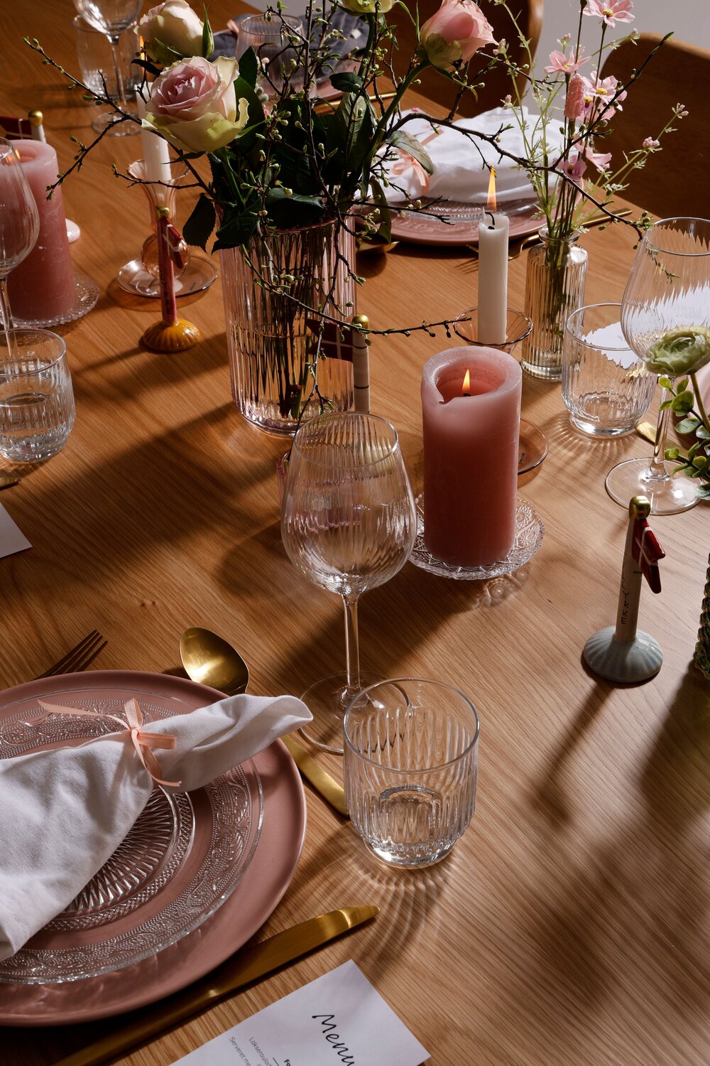 Konfirmationsborddækning i rosa nuancer på træbord. Bordet er dækket med en rosa tallerken, glastallerken, vinglas, vandglas, guld bestik og stofserviet. På midten af bordet er der pyntet med lyserøde bloklys, keramikflag, lysestager og vaser med blomster.