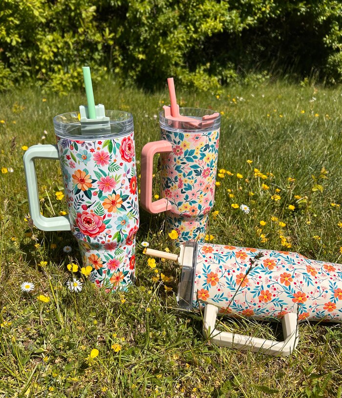 Store metal drikkedunke med sugerør med flotte sommerlige blomstermønstre ligger i græsset.