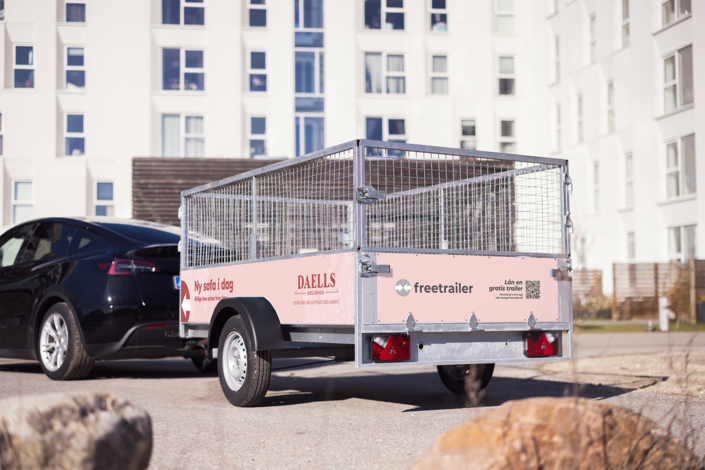 Lej en Freetrailer gittertrailer hos Daells Bolighus i Glostrup og Odense. Gittertraileren er hægtet på en sort bil foran et hvidt lejlighedskompleks.