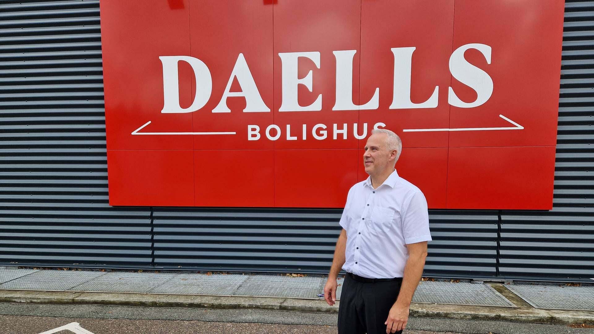 Brian Seeman Broe i hvid skjorte står foran en stor rød Daells logo med hvid tekst, mens den mørkgrå bygning danner baggrund.