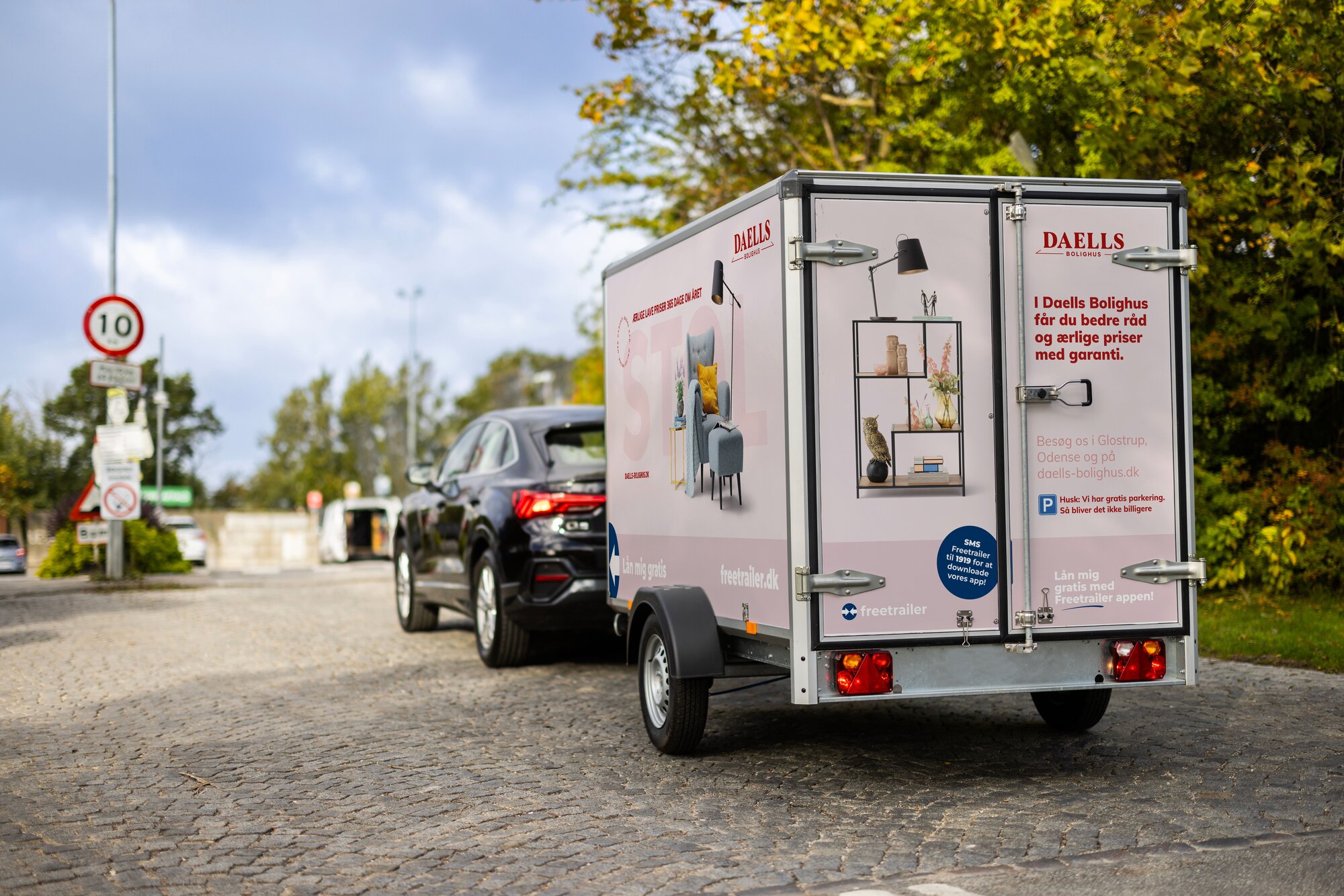 Daells Bolighus Freetrailer i lyserød bag sort bil på brostensvej med træer i baggrunden