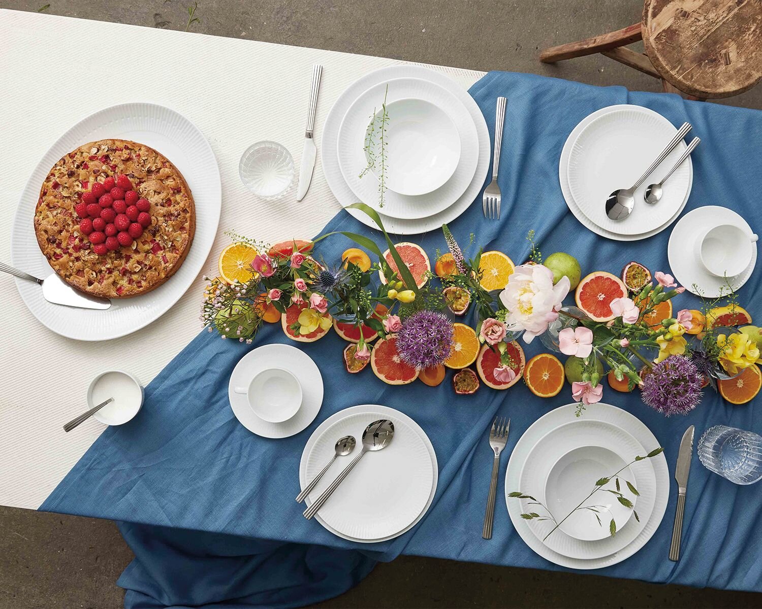 Farverigt dækket bord med hvidt stel, blå dug, friske blomster og citrusfrugter som bordpynt – inspiration til festlig borddækning fra Daells Bolighus.