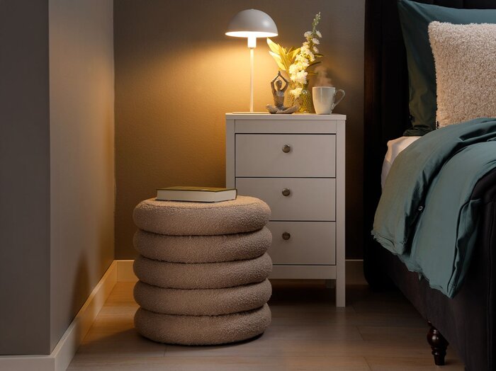 Rund puf med bløde i soveværelse med hvidt sengebord med skuffer og seng med blåt sengetøj.