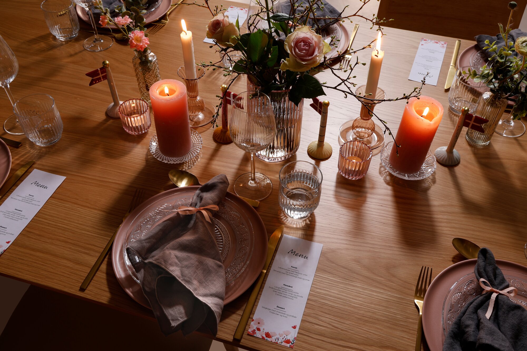 Borddækning til konfirmation med lyserøde nuancer. Bordet er dækket med rosa tallerkener, glas tallerkener, klare vinglas og vandglas. På midten er der pyntet med flag, lys og vaser med blomster.