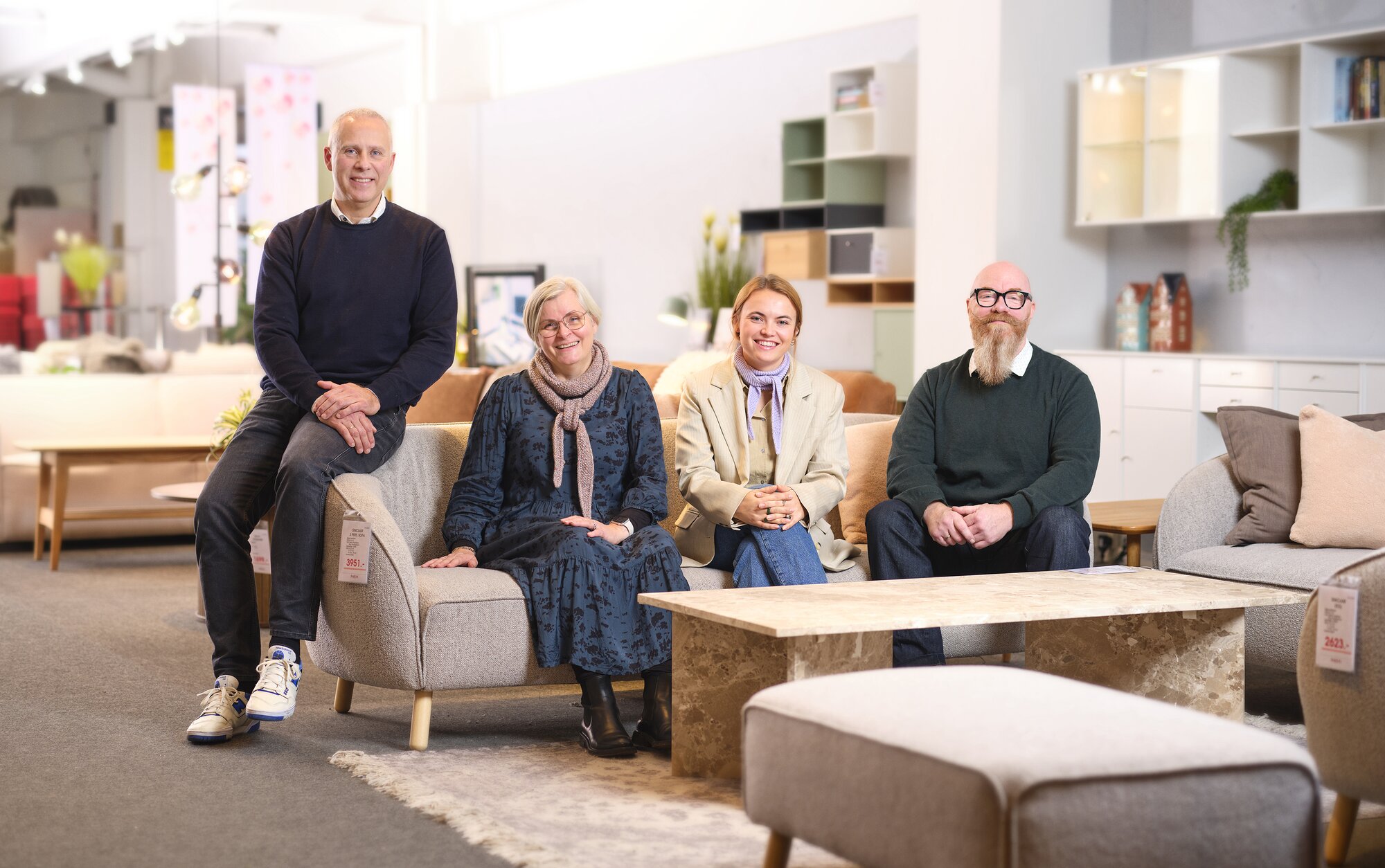 Fire personer sidder samlet på samme sofa i et lyst møbeludstillingsmiljø. Foran dem står et lavt sofabord, og bag dem ses udstillet boligindretning og hylder. De smiler og sidder i afslappede positurer.