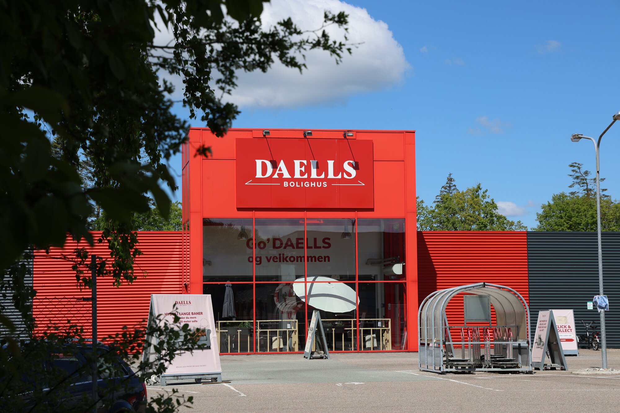 Rød butikfacade set udefra på en solrig dag med store vinduer, skiltning foran indgangen og et overdækket område til vogne på parkeringspladsen.