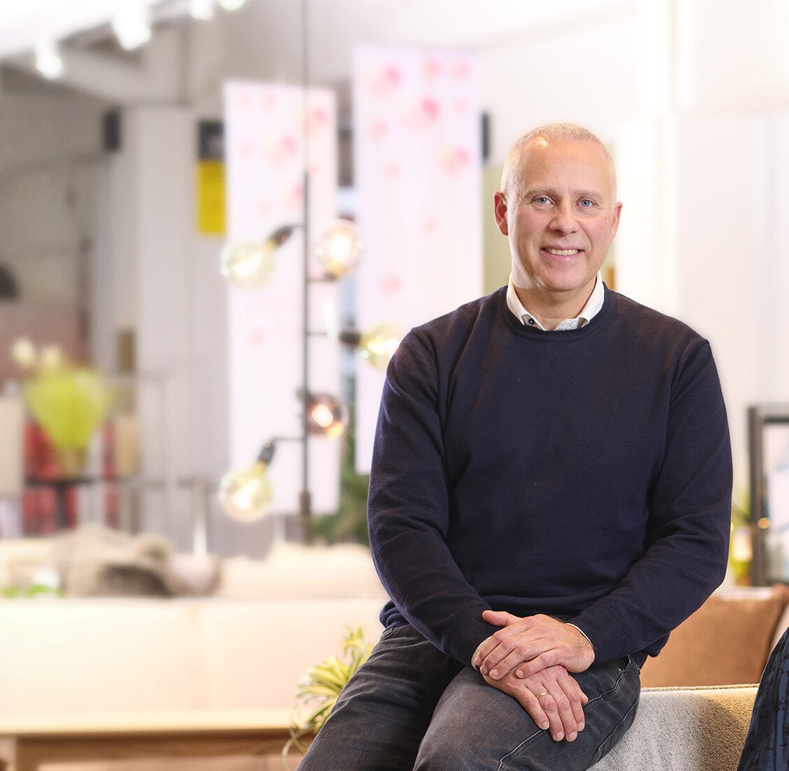 Brian Seemann Broe sidder afslappet i et lyst og moderne butiksmiljø med bløde møbler og dekorative lamper i baggrunden. Han smiler let og har hænderne foldet i skødet.