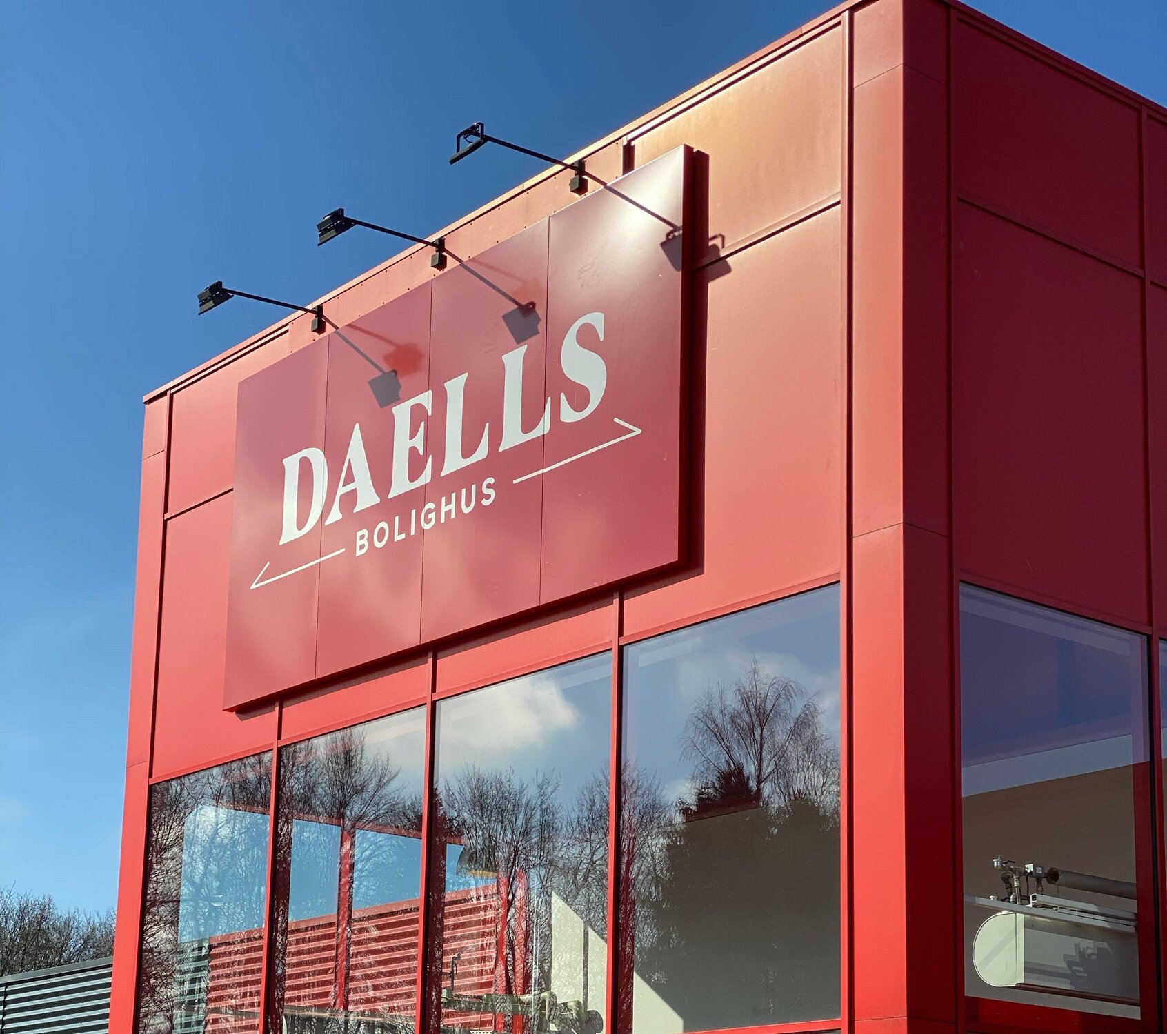 Rød butikfacade med stort hvidt skilt foroven og sollys der kaster refleksioner i de store vinduespartier. Bag vinduerne ses trætoppene spejle sig i glasset.