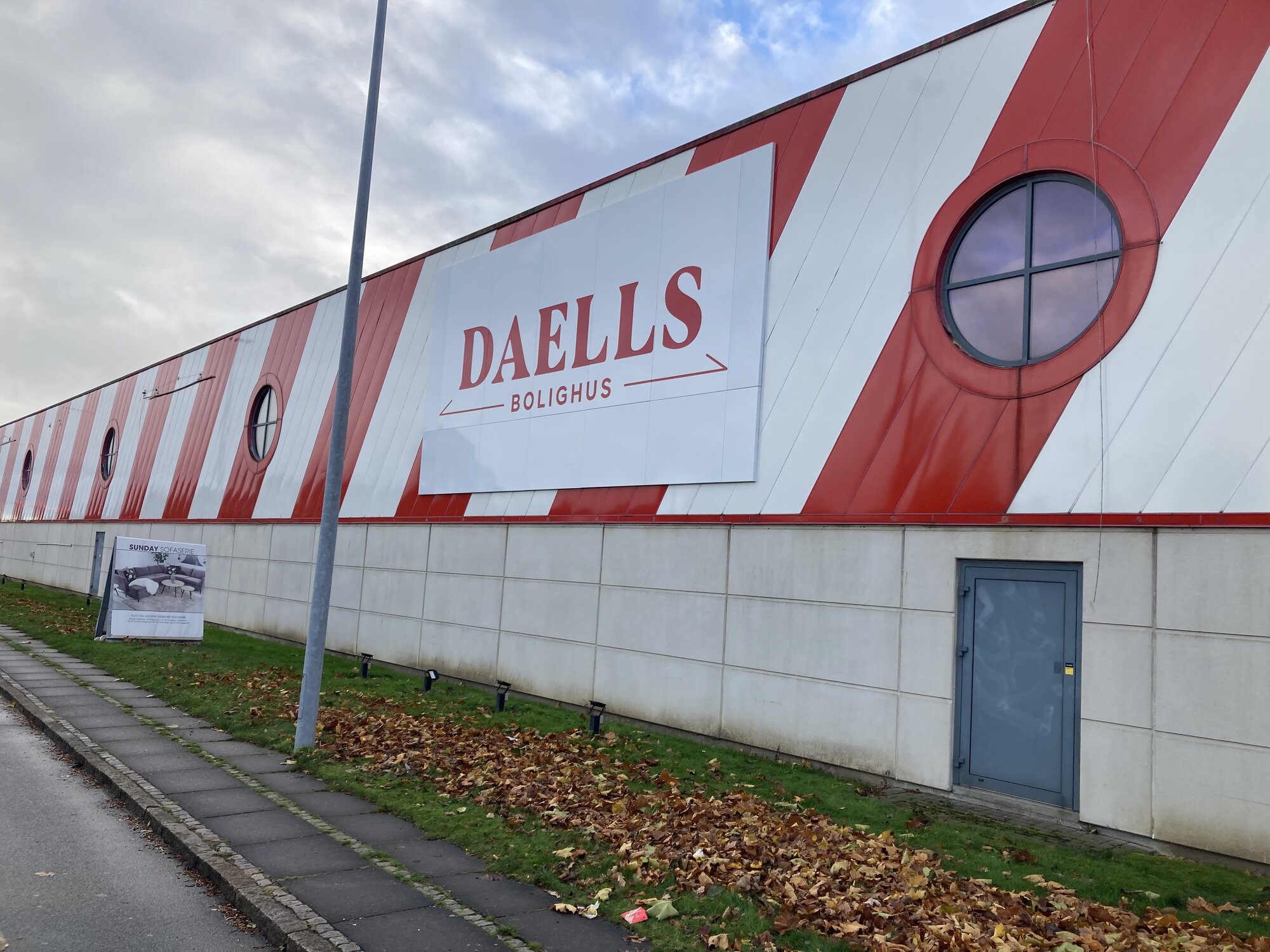 Lang bygning med skrå rød og hvid facade, runde vinduer og et stort skilt midt på væggen. Foran bygningen ligger en sti og en græsplæne dækket af efterårsblade under en grå himmel.