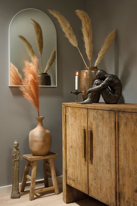 Boho indretning med stor massiv fyrretræs skænk, pyntet med keramikvase med pampasgræs, stor figur og en elegant lysestage. På væggen hænger et buet spejl, og på gulvet foran står en lille rustik træskammel med en keramik vase og ved siden af står en buddha figur.