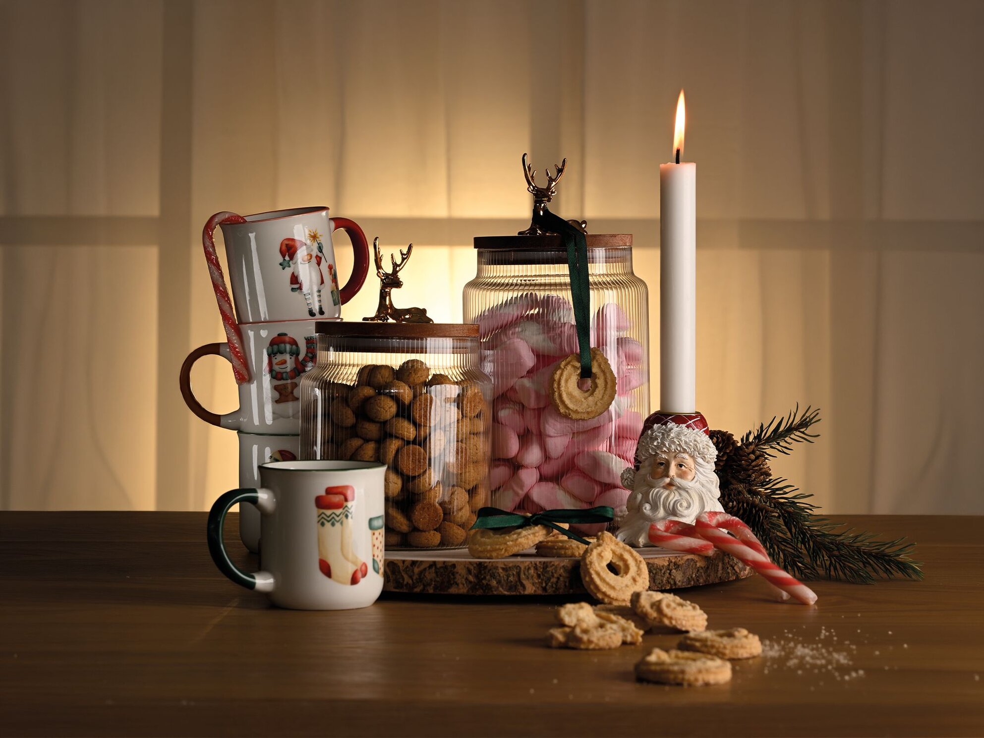 Stemningsfuldt julebord med julekrus med motiver, glaskrukker med pebernødder og juleskum, omringet af vaniljekranse, gran og en julemandslysestage.