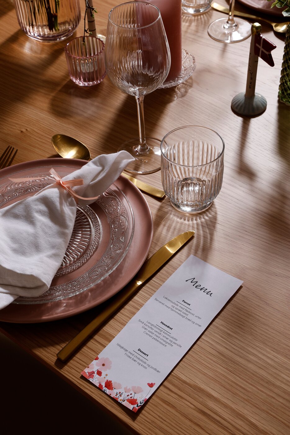 Træbord dækket med lyserød og glas tallerken. Klare vinglas og vandglas med mønster. Guld bestik og menu til gæsterne.