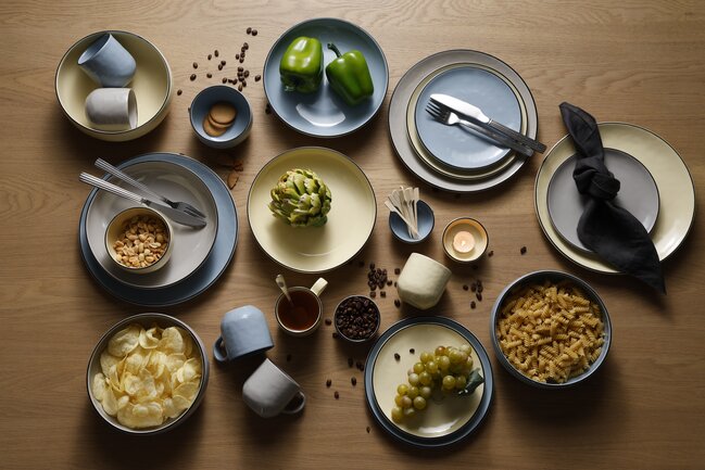 Træbord med gult, blåt og gråt Mandala stentøjsstel med sort kant. Bordet er fyldt med stellet, chips, kaffebønner, peanuts, vindruer, pasta, peberfrugt, småkager, tapaspinde, bestik og servietter.