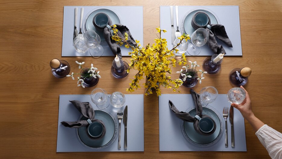 Påskebord dækket med blå dækkeservietter, blåt stel, og stofservietter. På midten af bordet står der blå stager med lys, blomster og æg. I midten står en forårsbuket med gule blomster. En arm rækker ind og stiller et vandglas på bordet.