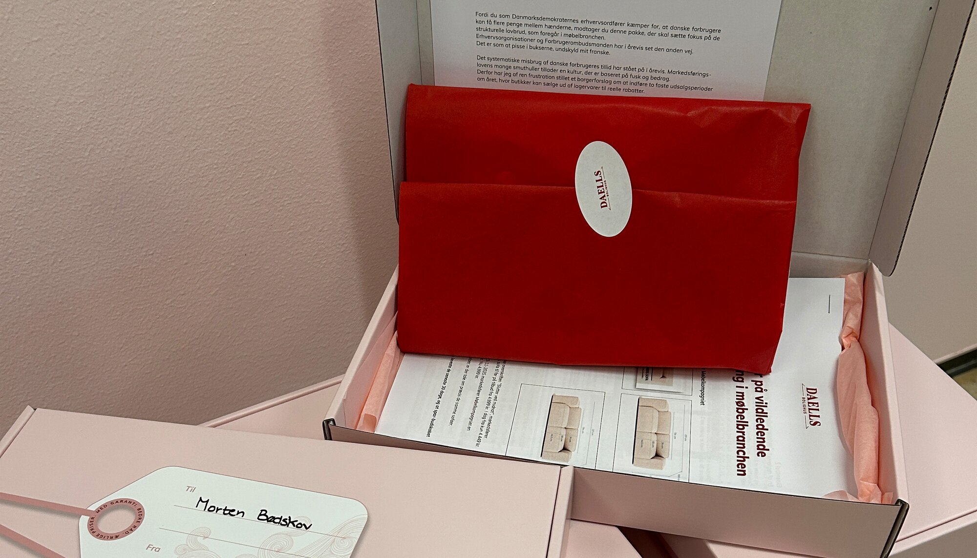 Åben gaveæske med lyserødt silkepapir, et rødt indpakket ble og papirer med tekst og illustrationer. Foran ligger en lukket lyserød æske med et til-og-fra-kort, hvor der står “Morten Bødskov”