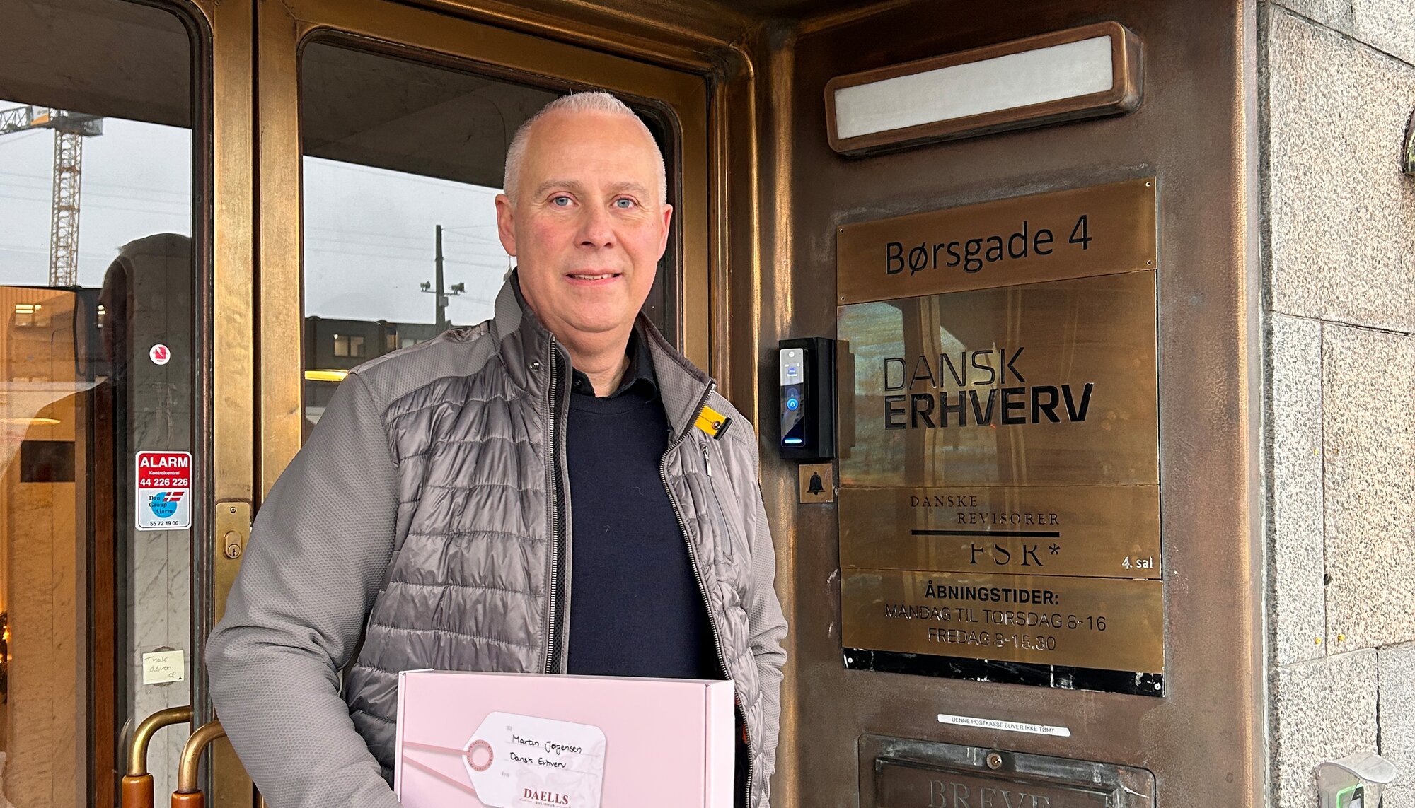 Mand står foran en indgang med messingdør og skiltning, hvor han holder en lyserød gaveæske med et til-og-fra-kort. Bagved ses bygningens adresse og åbningstider på en metalplade.